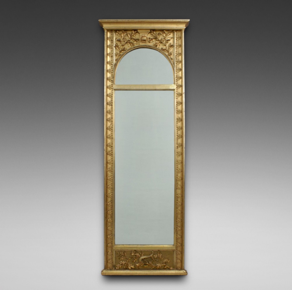 A tall, narrow early 19th-century French pier mirror featuring a gilded wood frame carved with oak leaves, an arched top plate, and a decorated lower plinth panel.