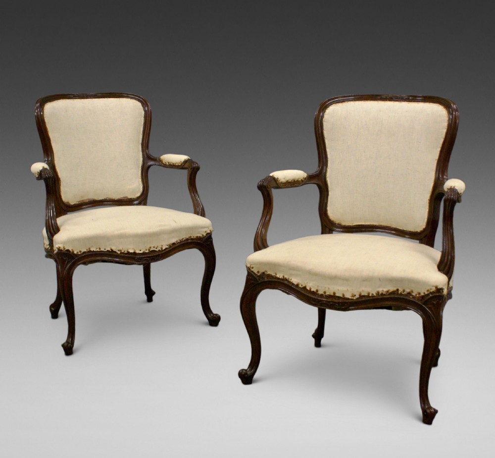 A pair of carved dark wood French Hepplewhite armchairs featuring cartouche backrests, padded armrests, and neutral cream fabric upholstery.