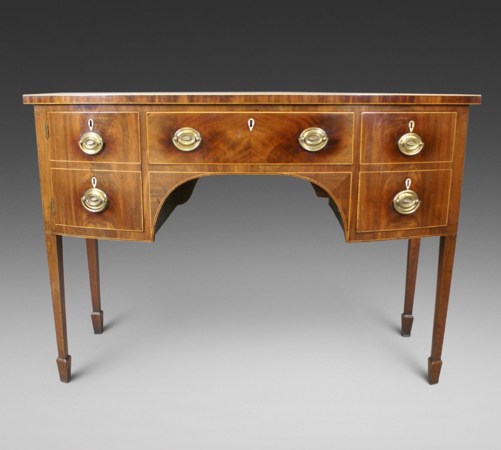 A George III mahogany sideboard featuring boxwood stringing, brass oval drop handles, a central arched apron, and tapered legs ending in spade feet.