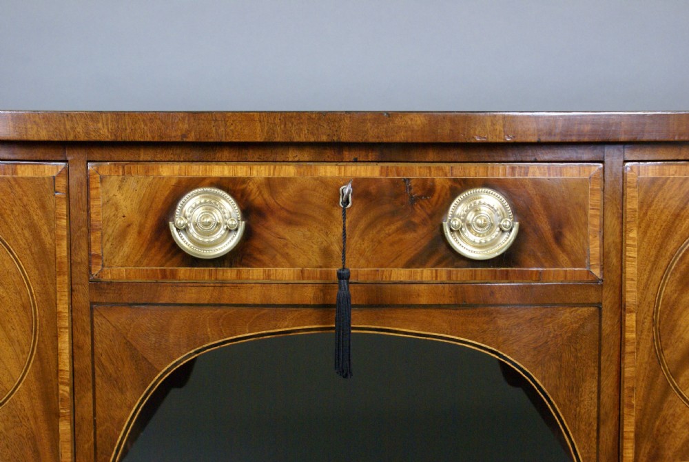 A George III flame mahogany sideboard featuring circular veneered door panels, boxwood stringing, brass circular ring handles, an arched central apron, and tapered square legs ending in spade feet. View IV