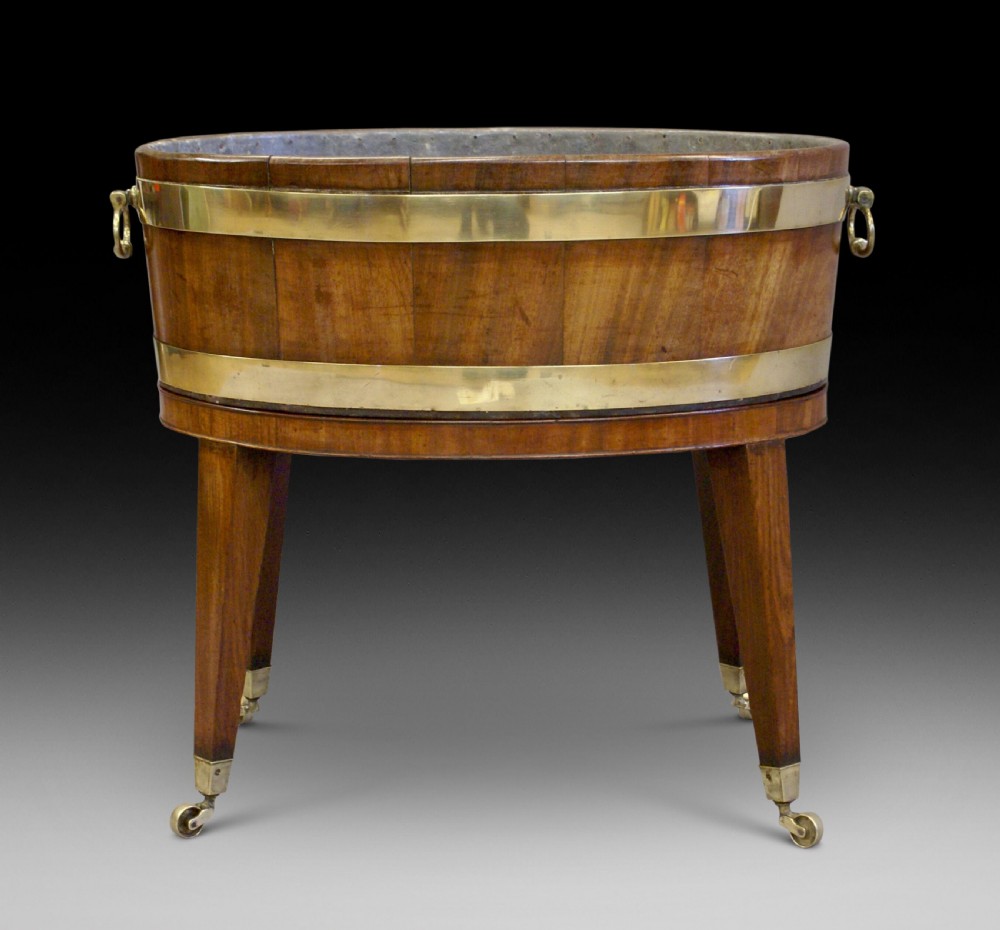 A George III oval mahogany wine cooler featuring brass binding hoops, twin handles, and tapered square legs on brass castors.