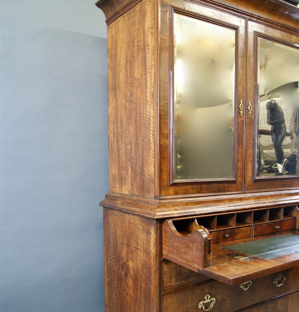 A George I figured walnut secretaire cabinet featuring a moulded upper section with two mirrored doors above a chest of drawers with brass drop handles. View VII