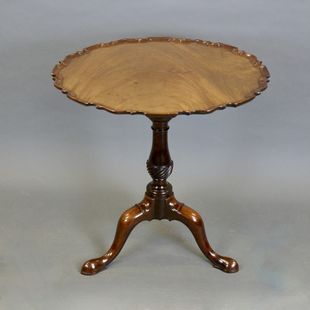 A George III mahogany tripod table displayed with its round piecrust top tilted vertically on a carved stem and cabriole legs. View II