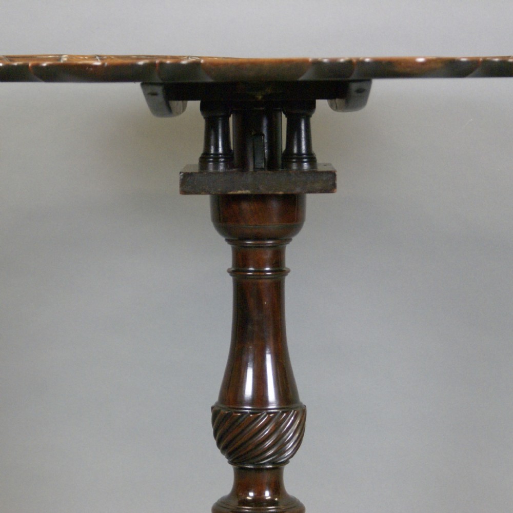 A George III mahogany tripod table displayed with its round piecrust top tilted vertically on a carved stem and cabriole legs. View III
