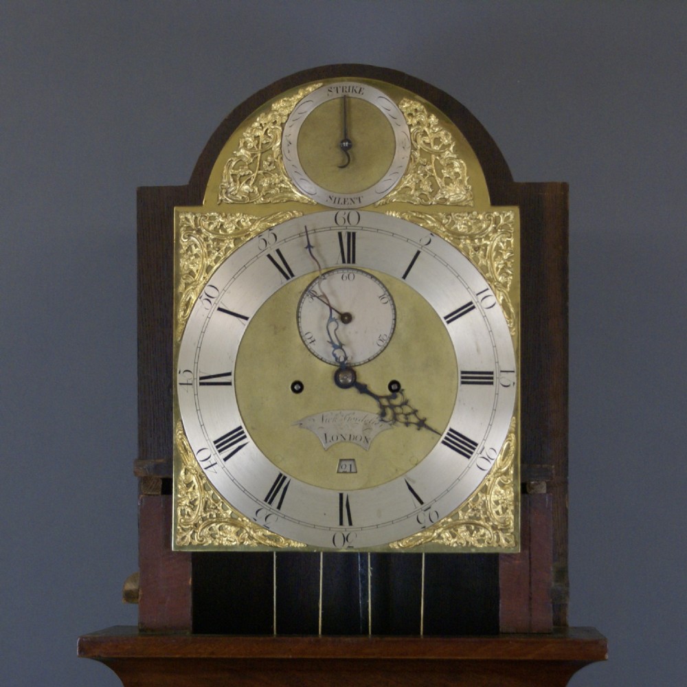 An antique Georgian mahogany longcase clock featuring an arched brass dial, fluted columns, eagle finials, and a flame mahogany trunk door. View VI