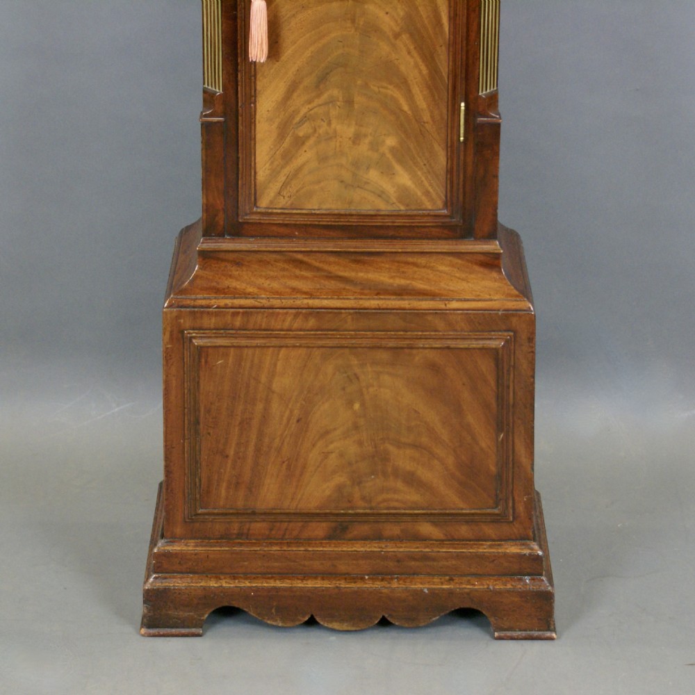 An antique Georgian mahogany longcase clock featuring an arched brass dial, fluted columns, eagle finials, and a flame mahogany trunk door. View VII