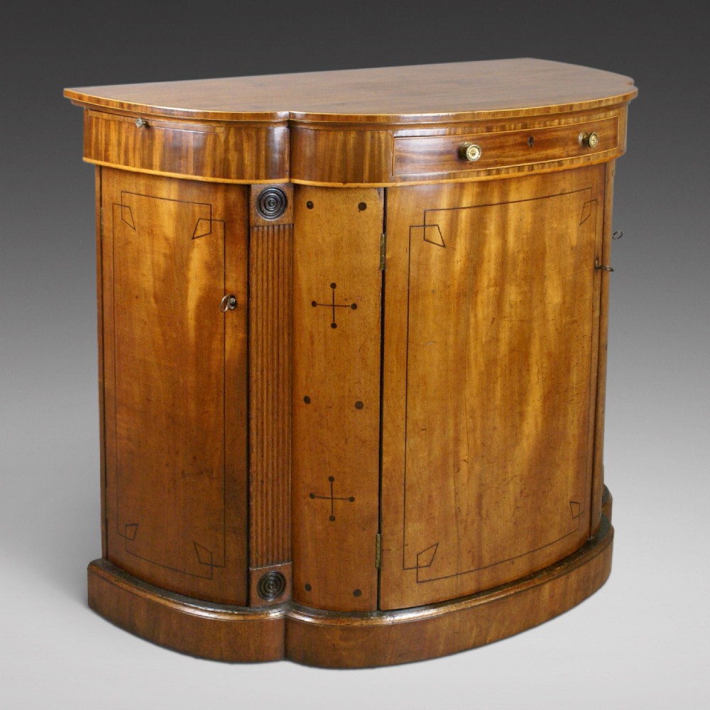An antique Regency mahogany side cabinet featuring a curved breakfront outline, ebony inlaid geometric details, fluted pilasters, and brass drawer knobs.
