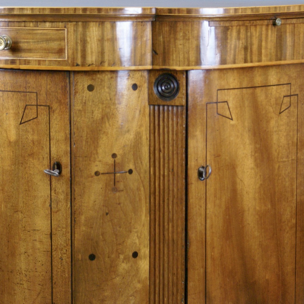 An antique Regency mahogany side cabinet featuring a curved breakfront outline, ebony inlaid geometric details, fluted pilasters, and brass drawer knobs. View V