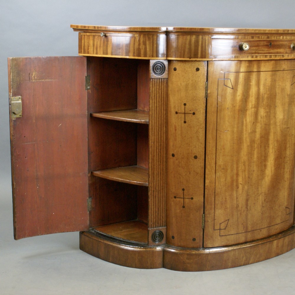 An antique Regency mahogany side cabinet featuring a curved breakfront outline, ebony inlaid geometric details, fluted pilasters, and brass drawer knobs. View VIII