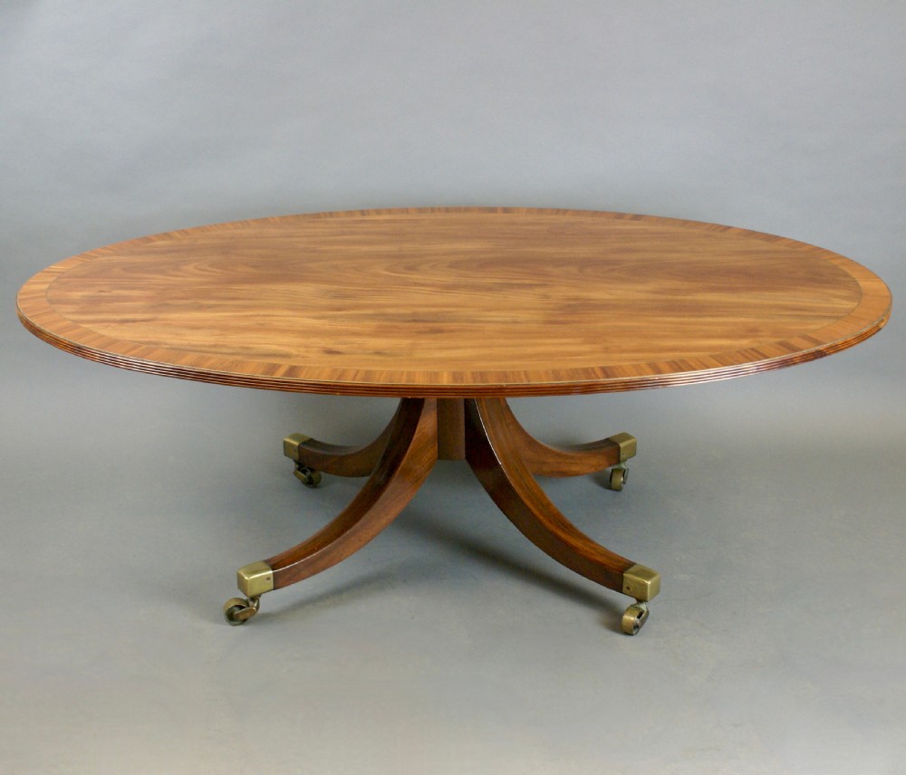 An oval flame mahogany coffee table with a crossbanded border resting on four splayed legs fitted with brass castors. View III