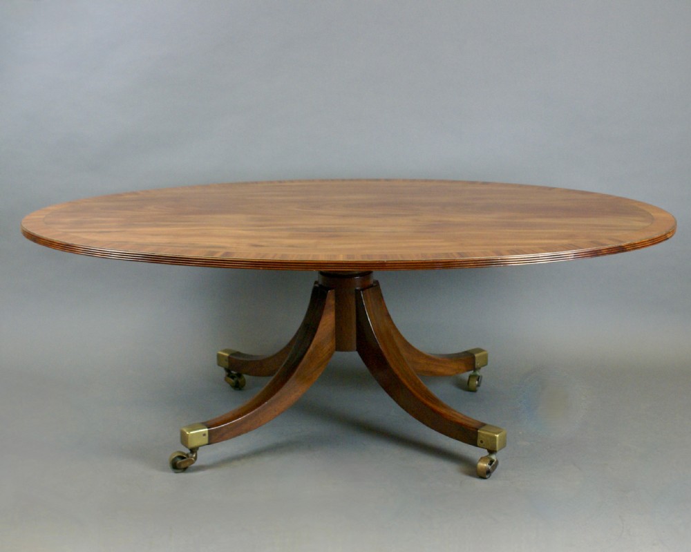 An oval flame mahogany coffee table with a crossbanded border resting on four splayed legs fitted with brass castors. View VI