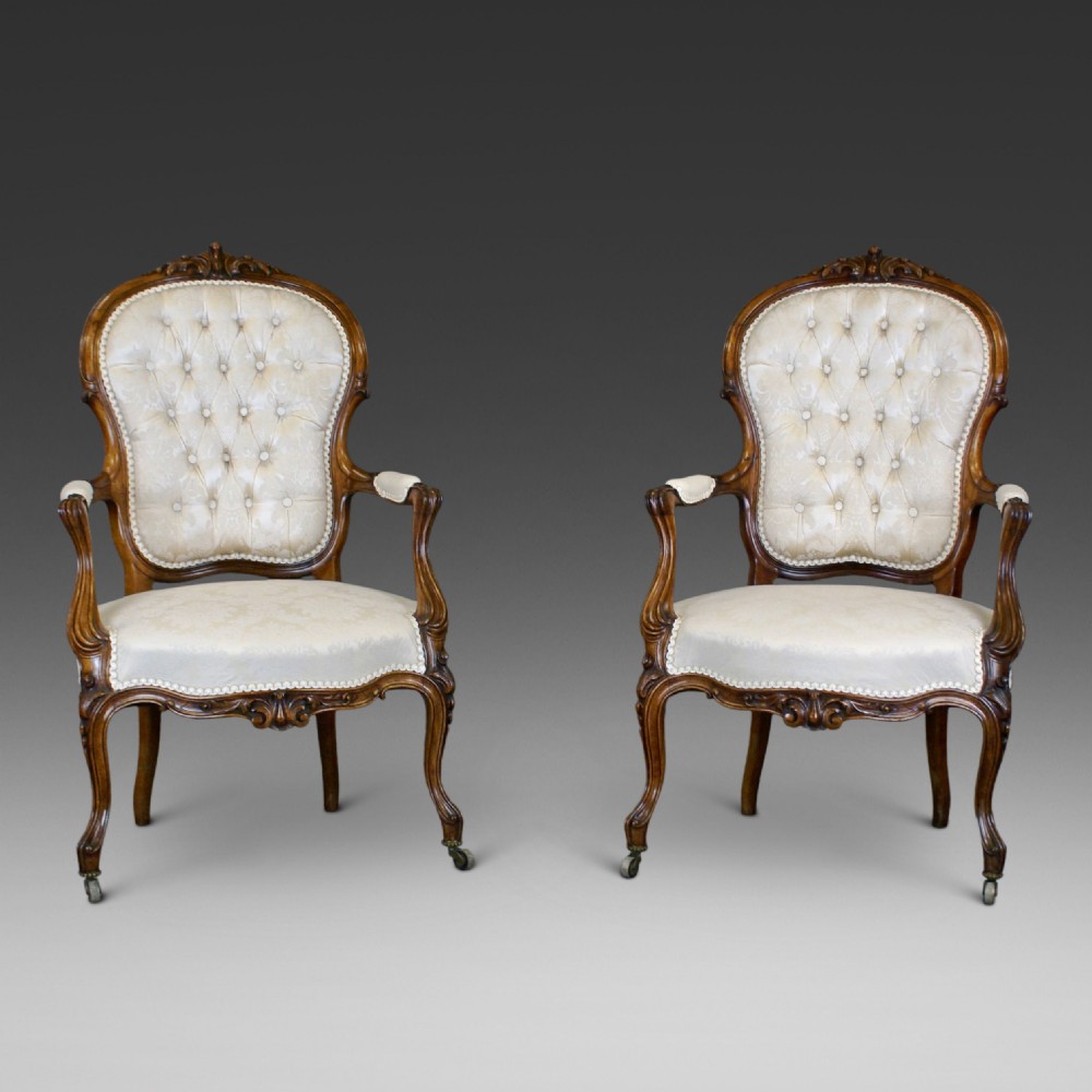 A pair of Victorian carved rosewood open armchairs with button-tufted cream upholstery and cabriole legs on brass castors.
