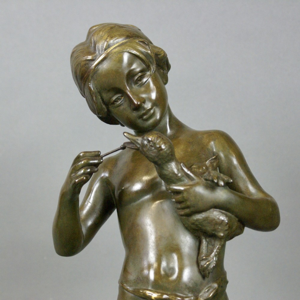 A patinated bronze figure depicting a standing young boy holding a small bird on a naturalistic cast base. View II