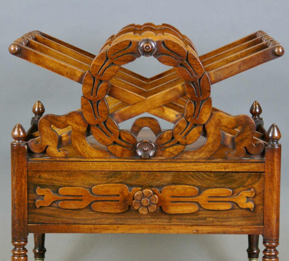 A Regency rosewood Canterbury featuring carved laurel wreath dividers, a single lower drawer with carved floral decoration, and turned legs on brass castors. View V