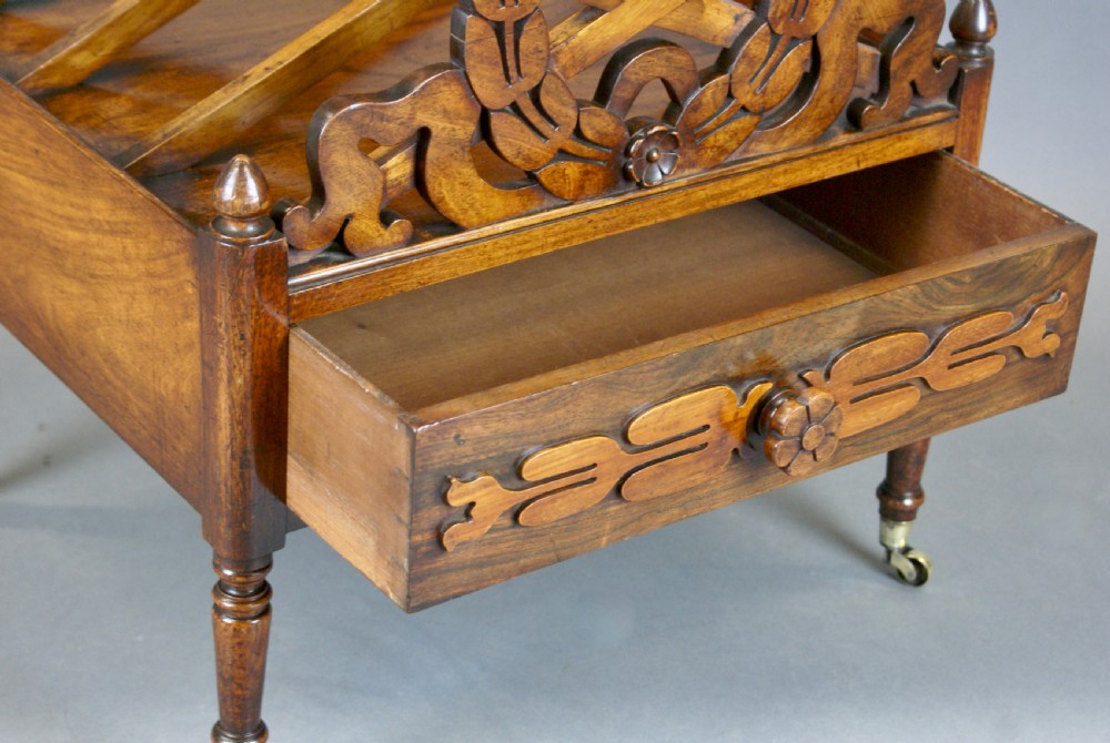 A Regency rosewood Canterbury featuring carved laurel wreath dividers, a single lower drawer with carved floral decoration, and turned legs on brass castors. View VI