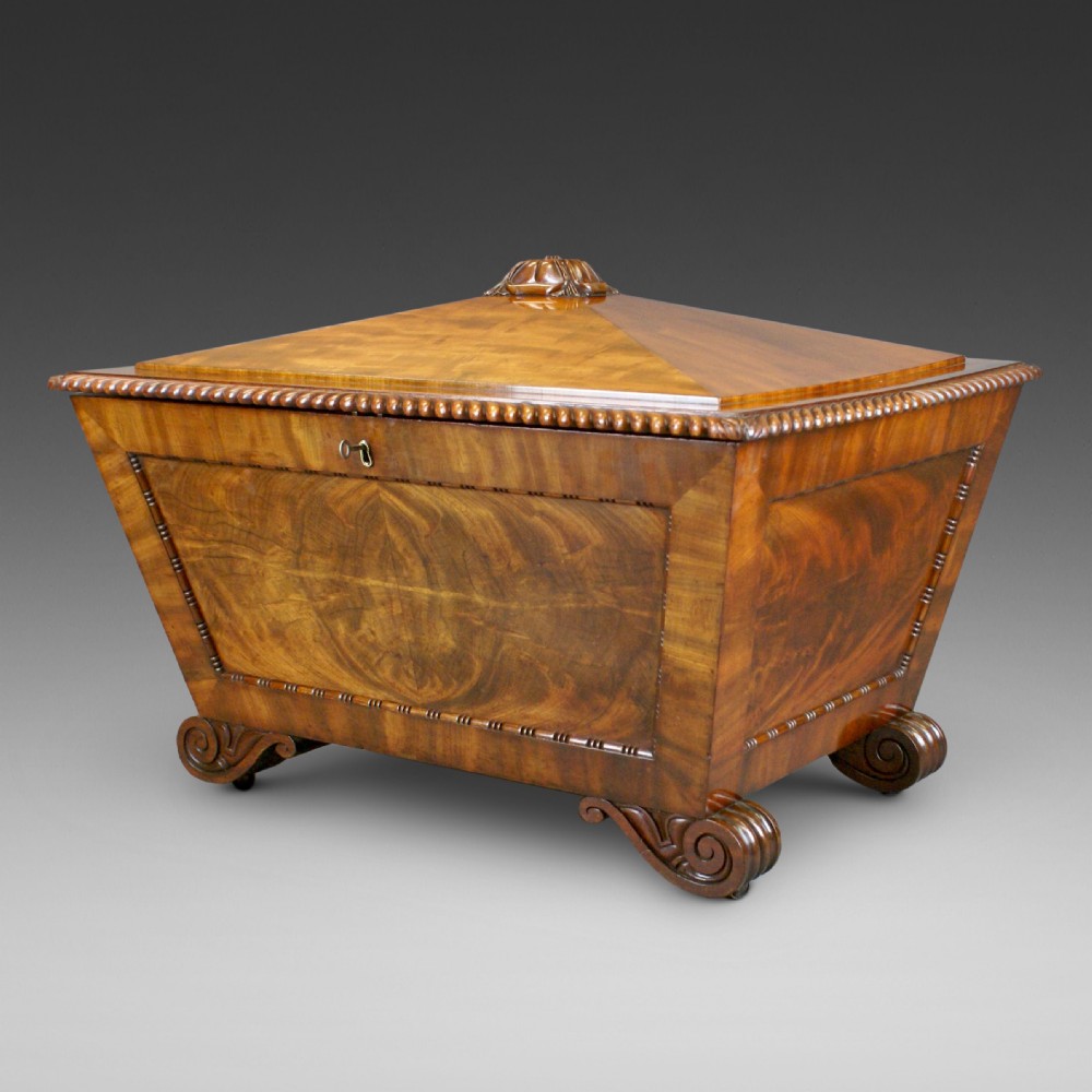 An antique Regency flame mahogany sarcophagus-shaped wine cooler featuring a pyramidal lid with carved finial, gadrooned borders, and scroll feet.