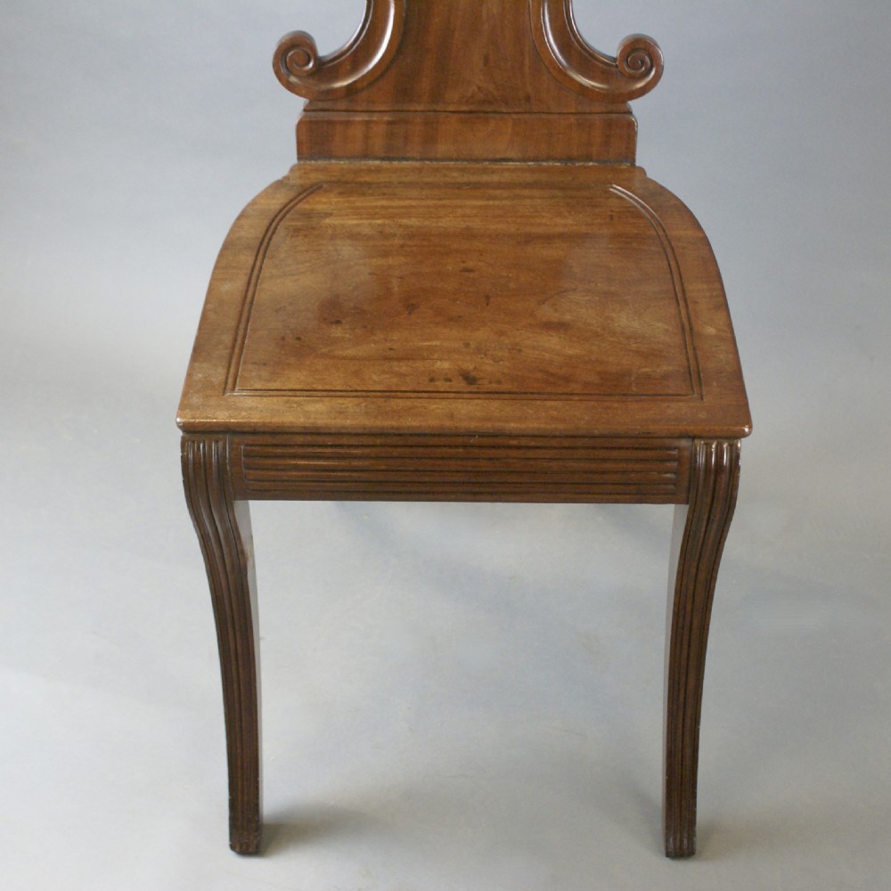 A pair of Regency mahogany hall chairs featuring carved cartouche backs with painted crest roundels, reeded seat rails, and sabre front legs. View VI