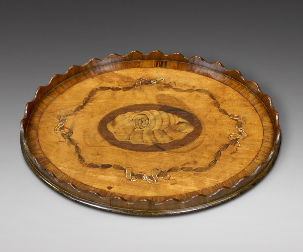 A miniature oval wooden tray featuring a central shell marquetry medallion surrounded by ribbon garland inlays and a wavy scalloped rim.