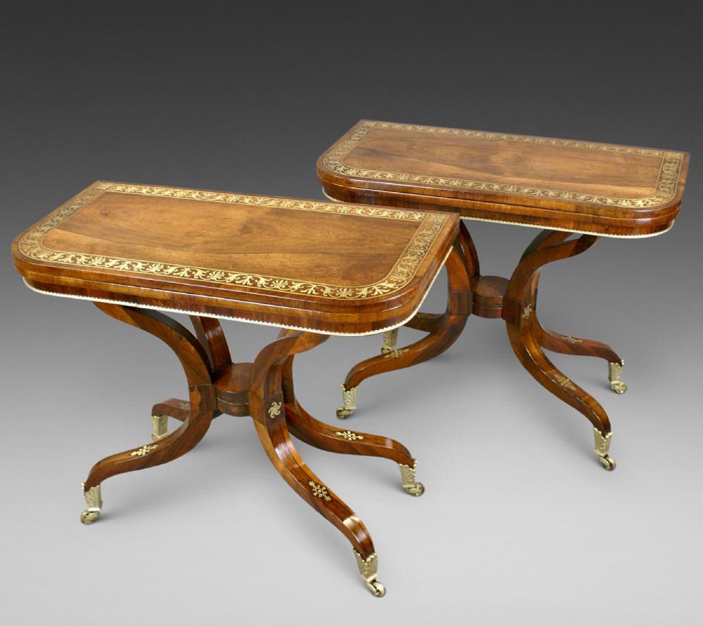 A pair of Regency dark rosewood card tables featuring fold-over rectangular tops decorated with brass floral inlays on curved sweeping legs with brass castors.