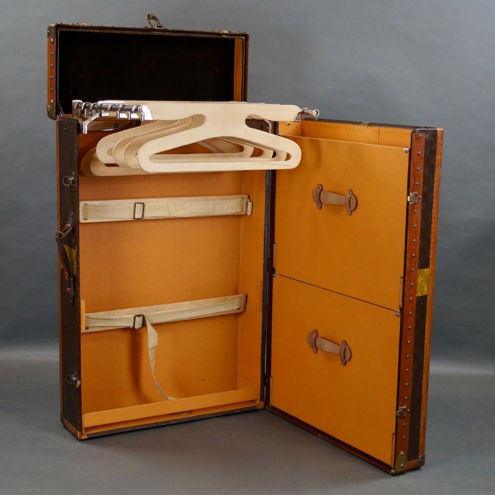 An upright dark brown monogram canvas trunk featuring a yellow painted cross band, leather edging, and brass latches. View III