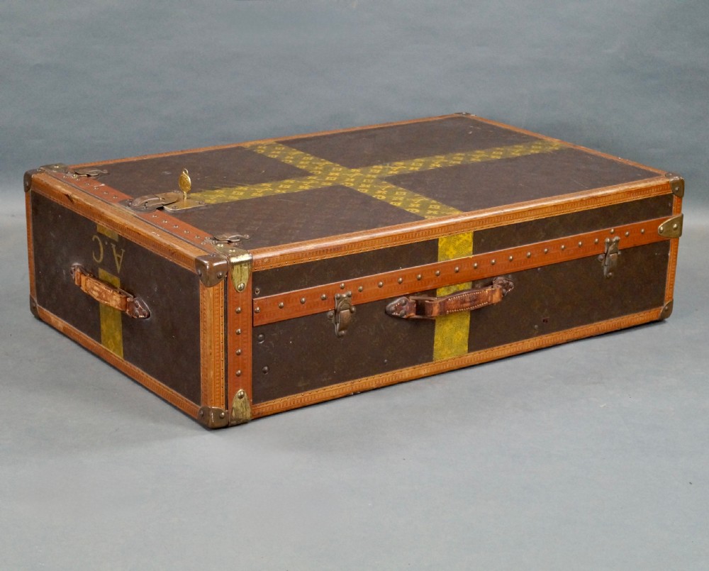An upright dark brown monogram canvas trunk featuring a yellow painted cross band, leather edging, and brass latches. View VII