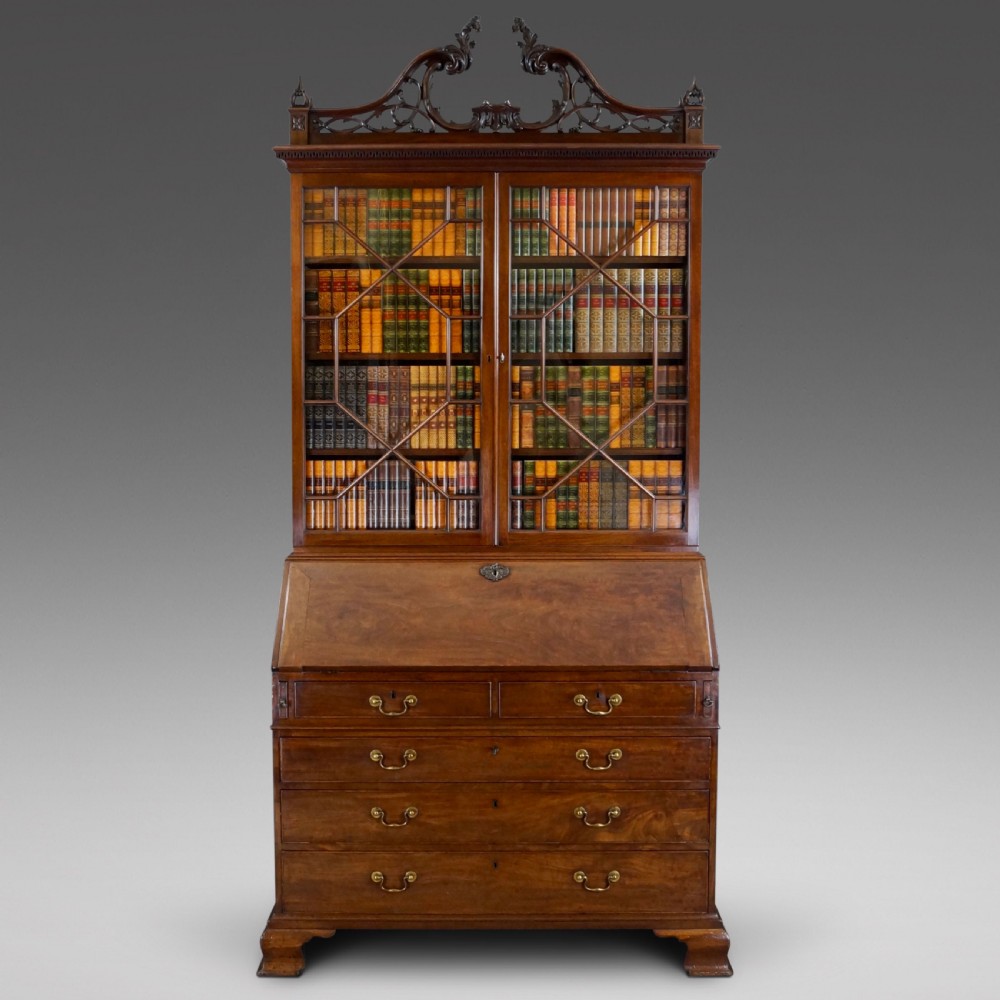 A George III mahogany bureau bookcase featuring a carved fretwork pediment, two astragal glazed upper doors, a sloping fall-front desk, and four graduated drawers with brass handles.