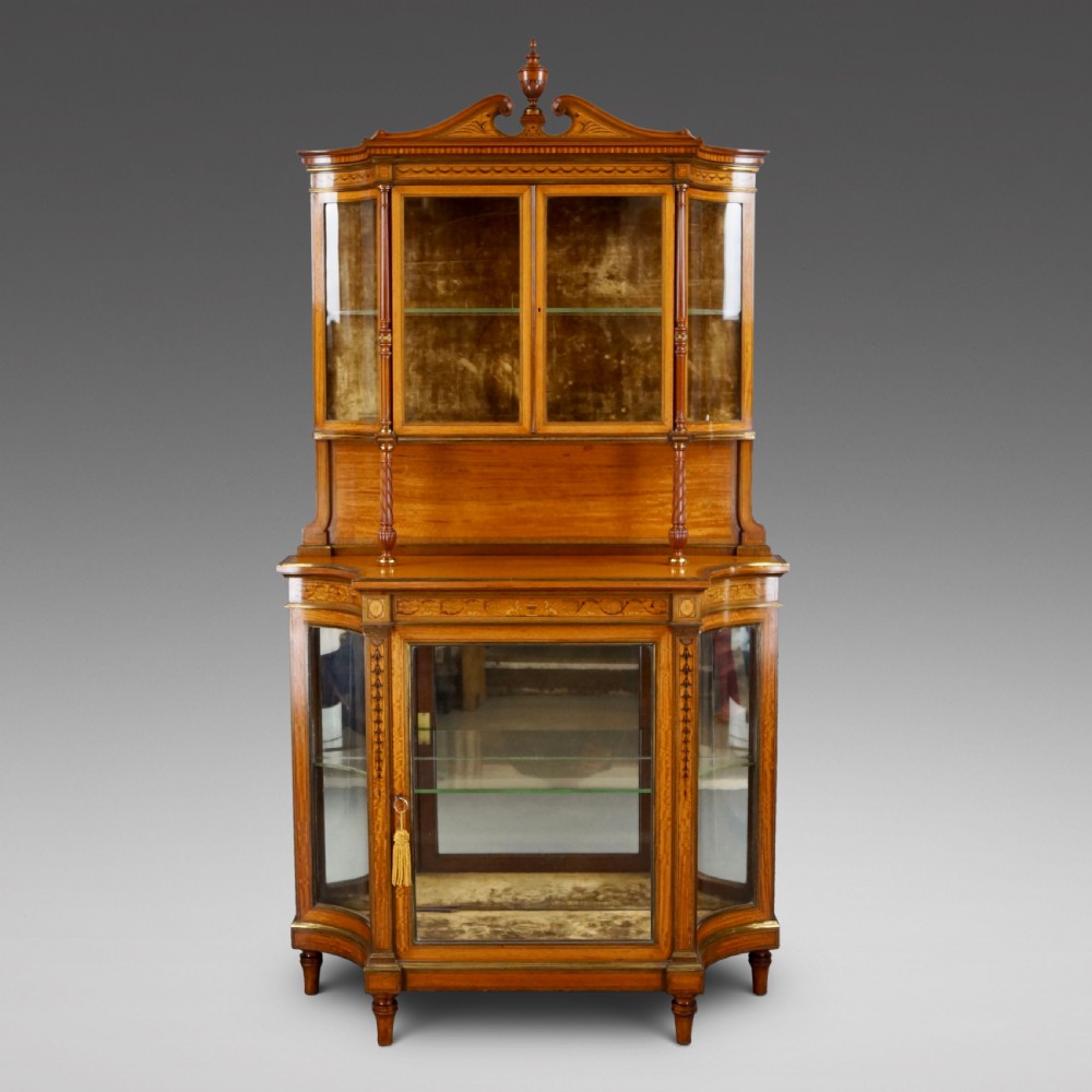An antique satinwood display cabinet featuring marquetry inlays, glazed double-tier doors, turned support columns, and an ornamental top finial.