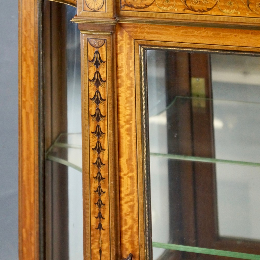 An antique satinwood display cabinet featuring marquetry inlays, glazed double-tier doors, turned support columns, and an ornamental top finial. View IV