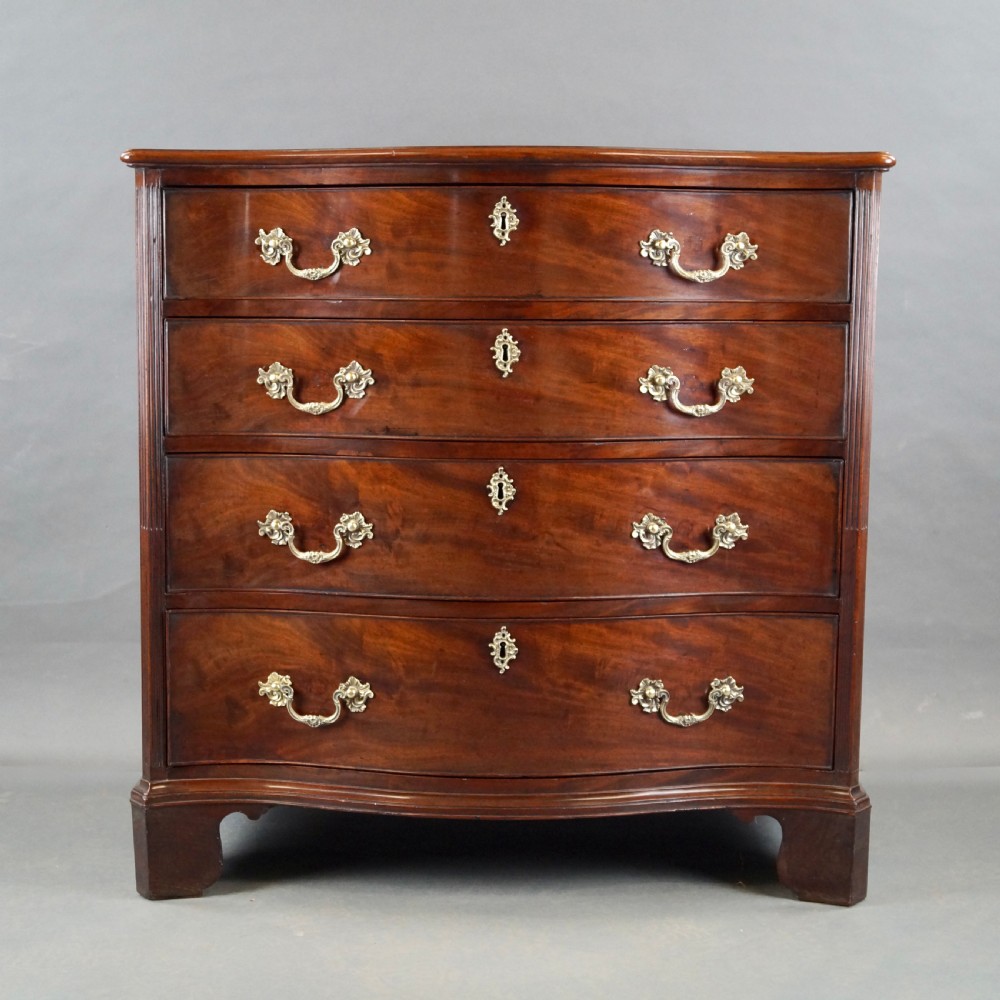 A George III flame mahogany serpentine chest of drawers featuring four graduated drawers with brass handles, keyhole escutcheons, bracket feet, and an oval inlaid top. View IV