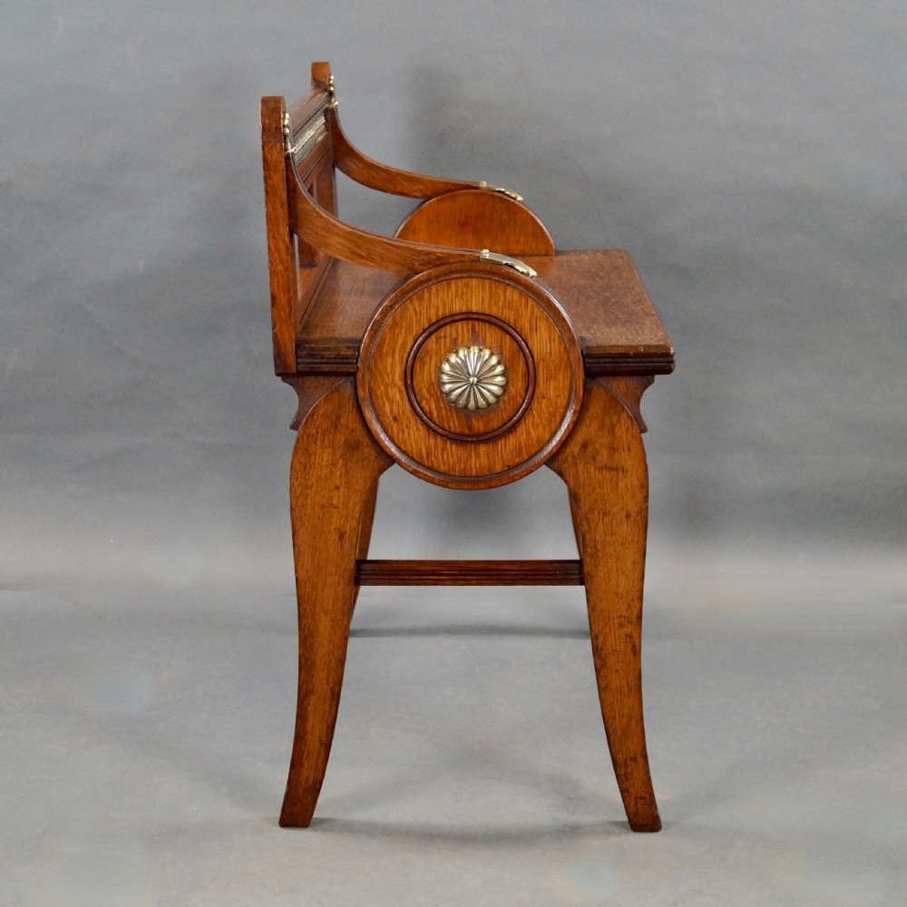 An oak hall seat featuring curved armrests with brass shell mounts, circular side roundels with central brass bosses, and a gallery-backed top rail. View III