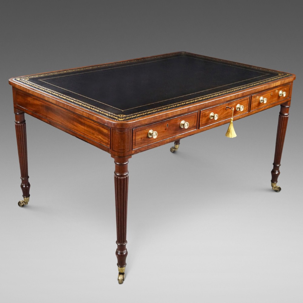 A George III mahogany partners writing table featuring a gold-tooled leather top, three drawers with brass knob handles, and fluted legs on brass castors.