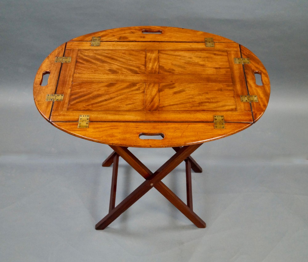 An antique oval flame mahogany butlers tray featuring brass flush hinges, cut-out handles, and a folding wooden stand with canvas webbing straps. View II