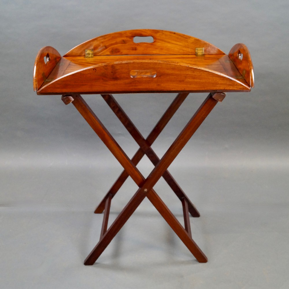 An antique oval flame mahogany butlers tray featuring brass flush hinges, cut-out handles, and a folding wooden stand with canvas webbing straps. View III