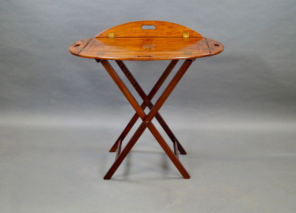 An antique oval flame mahogany butlers tray featuring brass flush hinges, cut-out handles, and a folding wooden stand with canvas webbing straps. View V