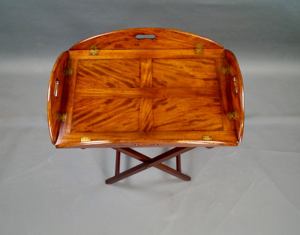 An antique oval flame mahogany butlers tray featuring brass flush hinges, cut-out handles, and a folding wooden stand with canvas webbing straps. View VI