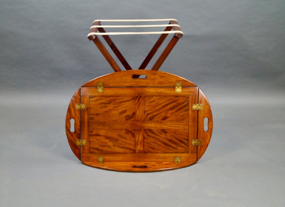 An antique oval flame mahogany butlers tray featuring brass flush hinges, cut-out handles, and a folding wooden stand with canvas webbing straps. View VII