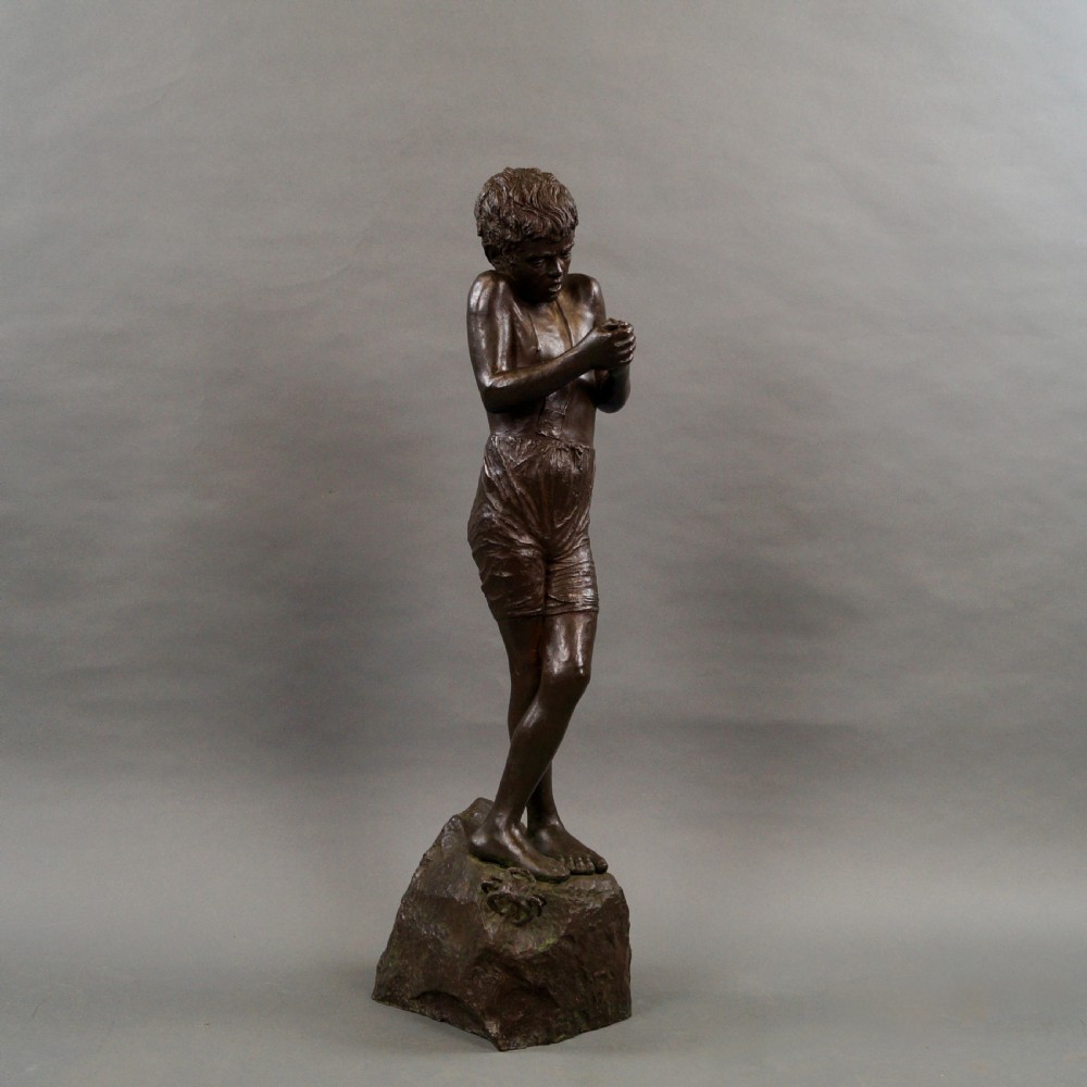 A dark brown bronze sculpture of a young boy standing with crossed legs on a textured rock base, holding his hands together. View III