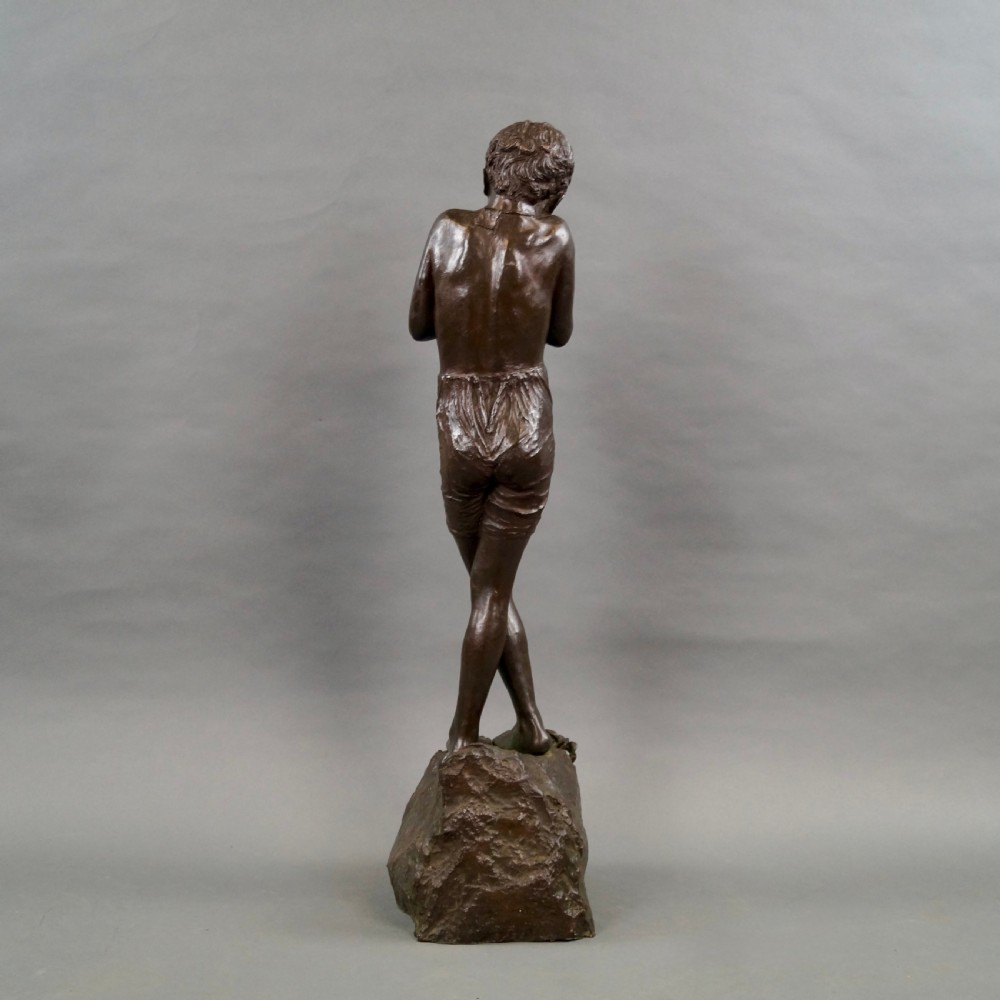 A dark brown bronze sculpture of a young boy standing with crossed legs on a textured rock base, holding his hands together. View V
