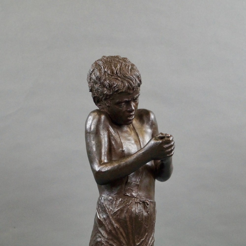 A dark brown bronze sculpture of a young boy standing with crossed legs on a textured rock base, holding his hands together. View VI
