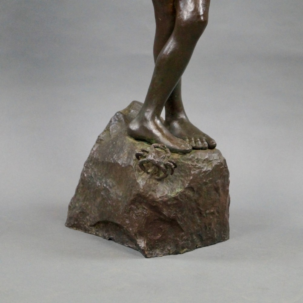 A dark brown bronze sculpture of a young boy standing with crossed legs on a textured rock base, holding his hands together. View VII