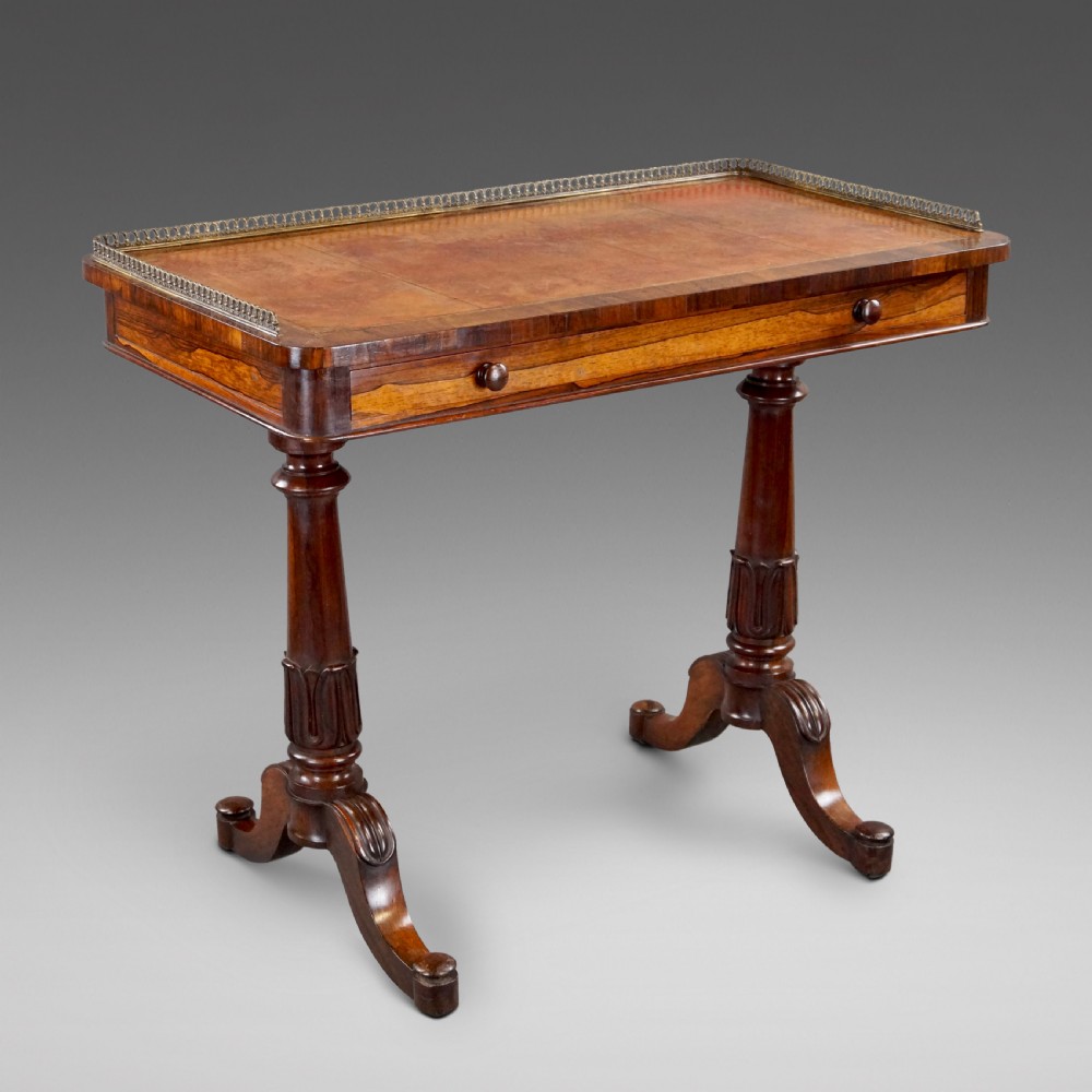 A Victorian rosewood writing table featuring a brass gallery rail around three edges, a front drawer with turned wooden knobs, and twin carved pedestal legs.