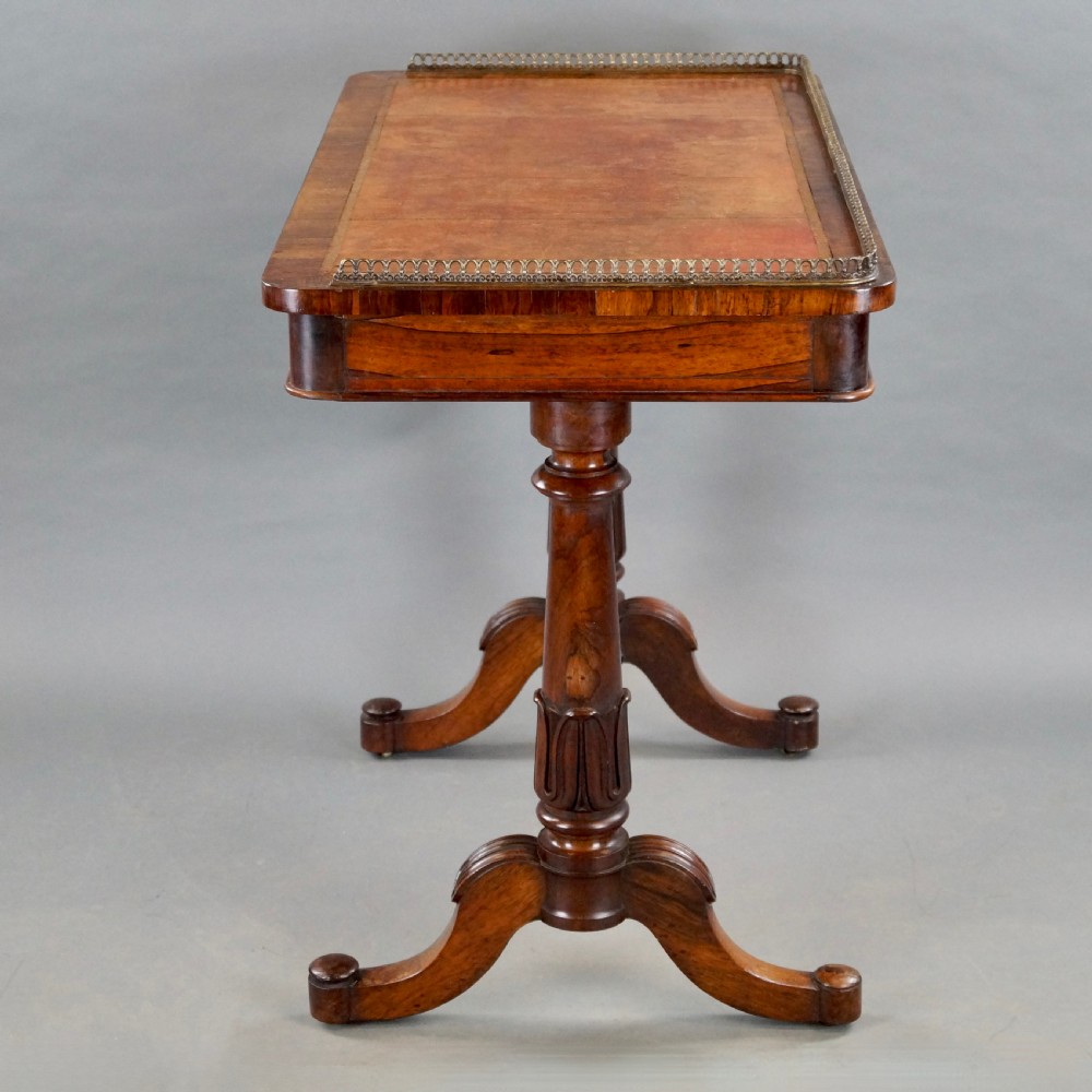 A Victorian rosewood writing table featuring a brass gallery rail around three edges, a front drawer with turned wooden knobs, and twin carved pedestal legs. View III