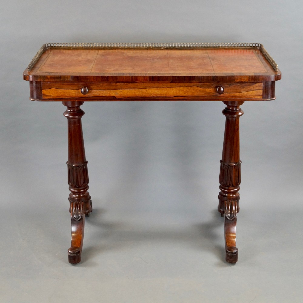 A Victorian rosewood writing table featuring a brass gallery rail around three edges, a front drawer with turned wooden knobs, and twin carved pedestal legs. View VI