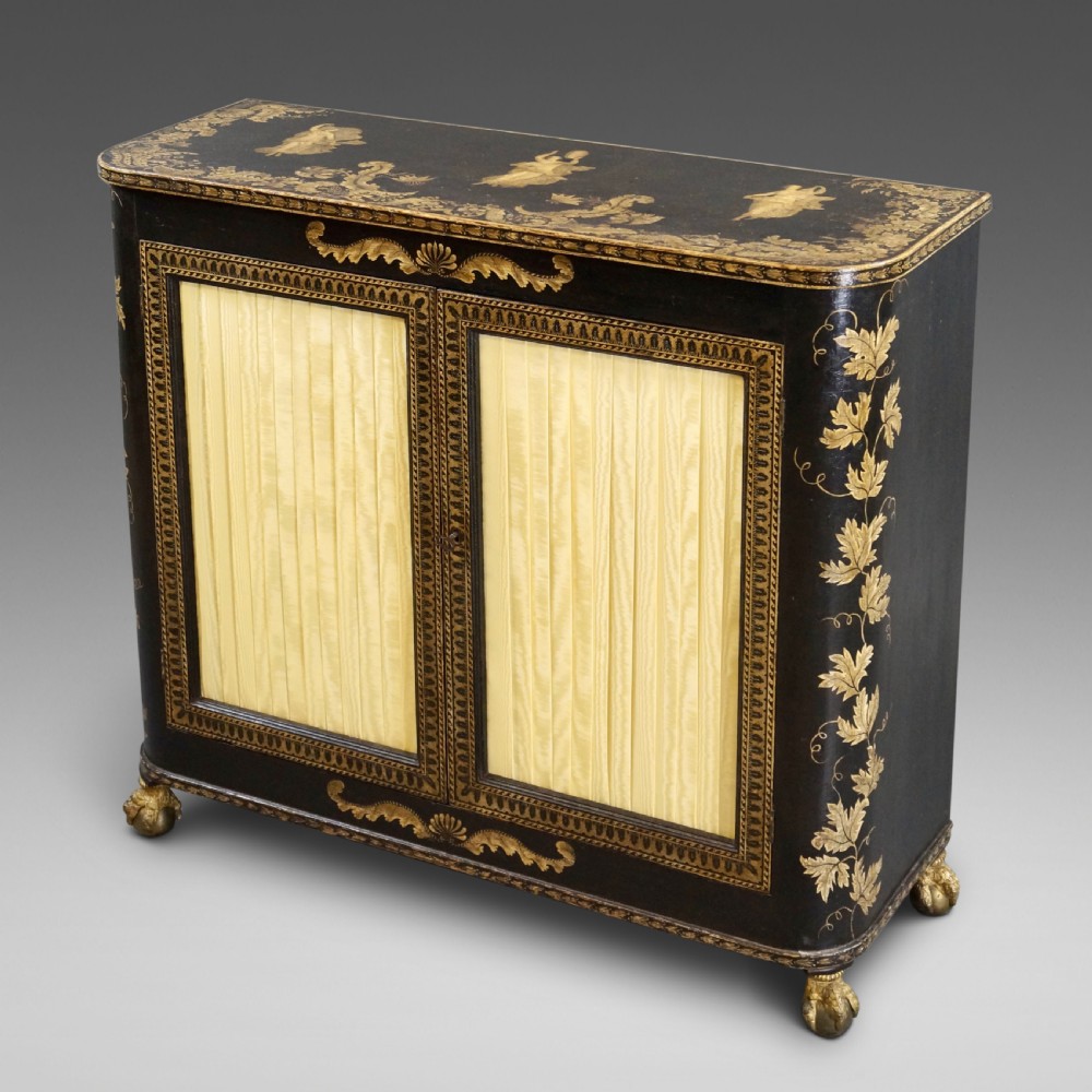 A Regency period black pen-work side cabinet featuring gold vine motifs, pleated yellow silk door panels, and gilded paw feet.