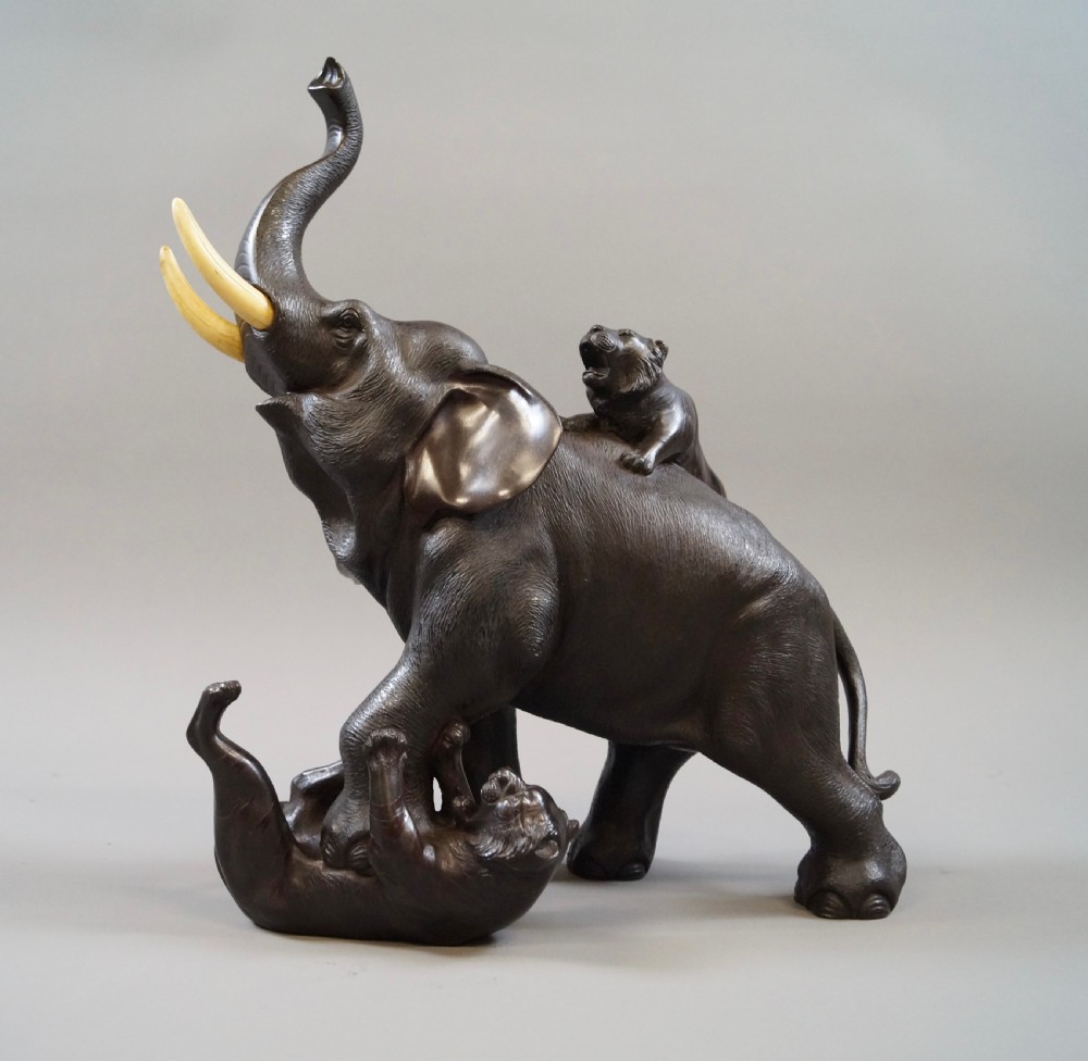 A dark brown patinated bronze okimono sculpture depicting an elephant fighting two tigers, featuring inlaid light tusks.