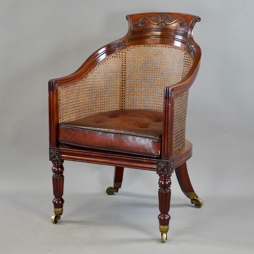 A Regency mahogany bergere armchair with a carved cresting rail, woven cane back and sides, a buttoned brown leather seat cushion, and brass castors.