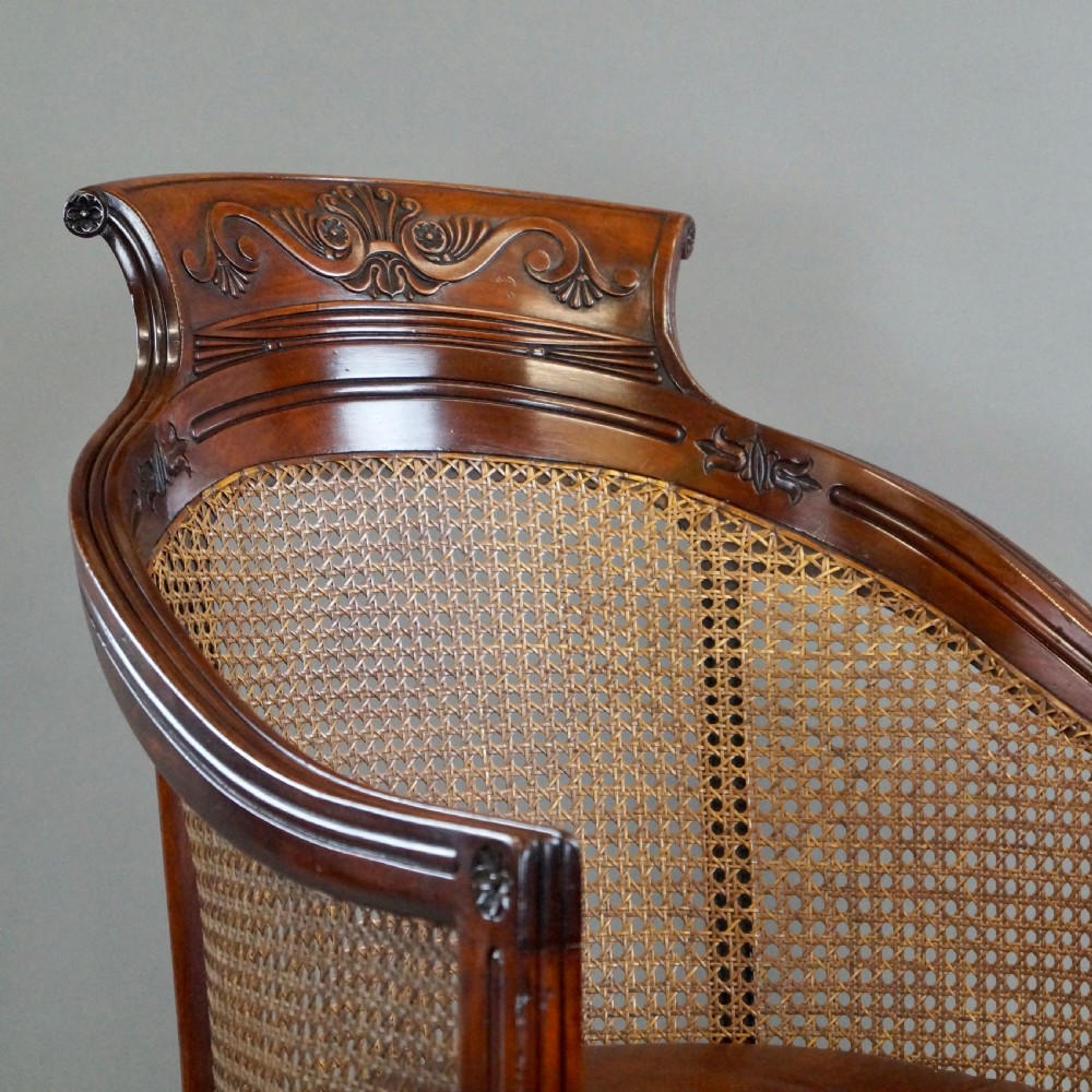 A Regency mahogany bergere armchair with a carved cresting rail, woven cane back and sides, a buttoned brown leather seat cushion, and brass castors. View II