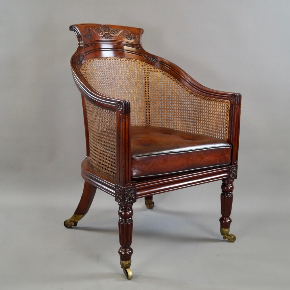 A Regency mahogany bergere armchair with a carved cresting rail, woven cane back and sides, a buttoned brown leather seat cushion, and brass castors. View III