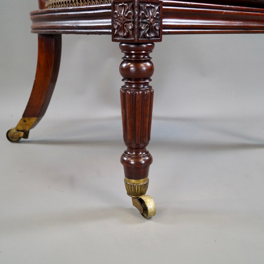 A Regency mahogany bergere armchair with a carved cresting rail, woven cane back and sides, a buttoned brown leather seat cushion, and brass castors. View V