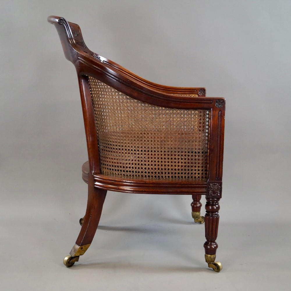 A Regency mahogany bergere armchair with a carved cresting rail, woven cane back and sides, a buttoned brown leather seat cushion, and brass castors. View VI